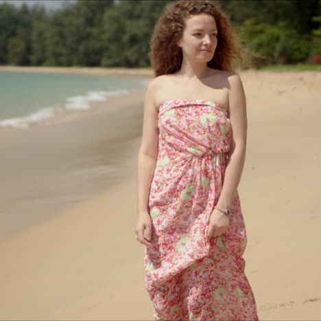 Pleasant-woman-holding-flowered-dress-and-walking-on-sandy-beach-in-bright-day