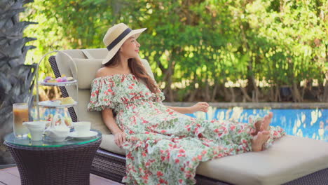 a young asian woman in a long loose dress and straw sun hat reclines poolside for tea time