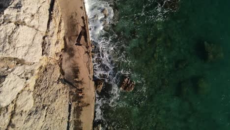 Take-a-floating-aeria-with-drone-on-a-woman-who-to-her-right-is-the-sea-and-to-her-left-a-great-mountain-you-can-see-the-rocks-on-the-coast-and-its-transparent-water-great-shot-4k