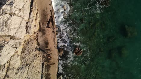 toma un aeria flotante con drone sobre una mujer que a su derecha esta el mar y a su izquierda una gran montaña se ven las rocas en la costa y su agua transparente gran toma 4k