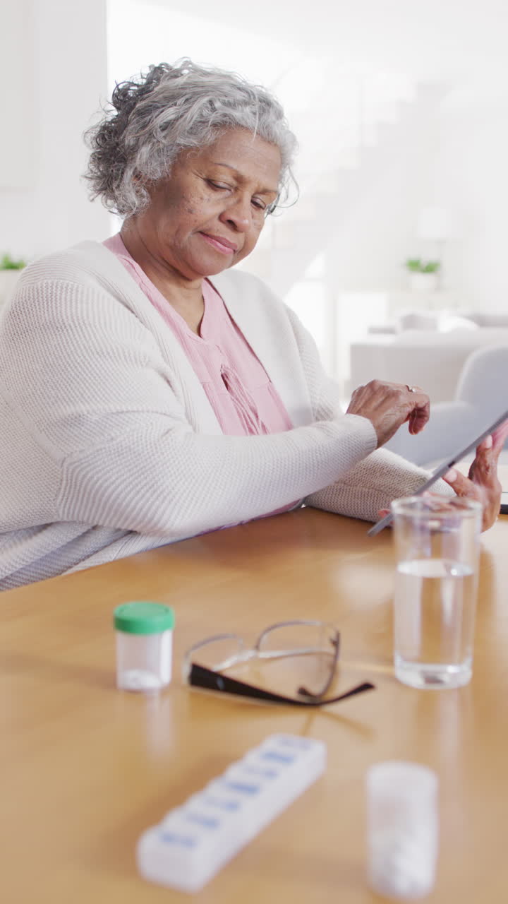 Vertical video of senior african american woman sitting at table, taking pills