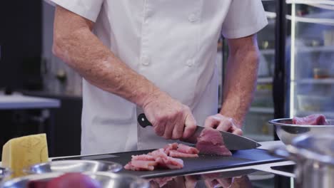 cocinero caucásico vestido de blanco en una cocina de restaurante cortando carne en una tabla de cortar