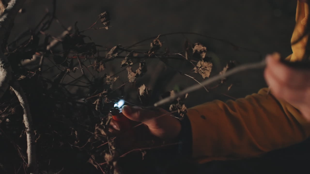 Kvinde tænder tørre kviste på en campingplads ved søen, flammen vokser mellem grenene, varm orange belysning og røg, nærbillede af hånd der holder tændstik, hyggelig natlig komfort og tilfredshed.