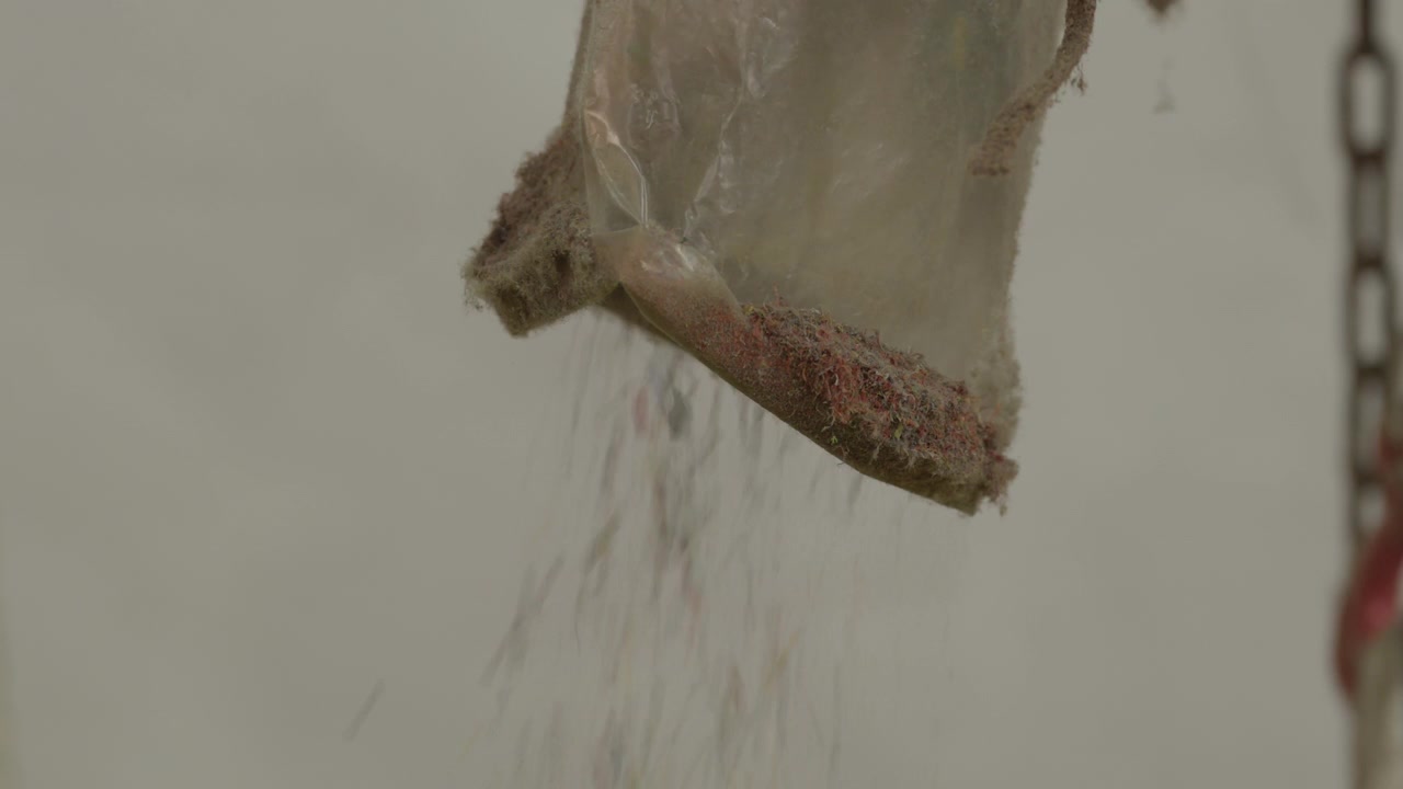 Granuli di plastica bianchi fuoriescono dal fondo strappato di un saccone sospeso da una catena in una fabbrica di riciclaggio. Primo piano.