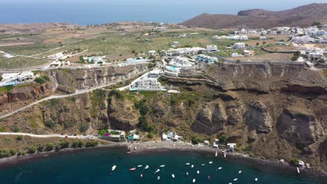 Aerial-flying-towards-Greek-Island,-rocky-cliff-with-villas-and-fishing-boats-in-the-Mediterranean-Sea-on-beach-in-Santorini,-Greece