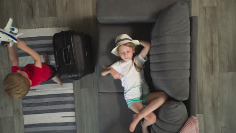 tired girl waves hand lying on sofa while toddler playing