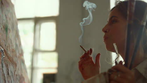 Female-painter-blowing-smoke-out-indoors.-Creative-girl-looking-to-artwork.