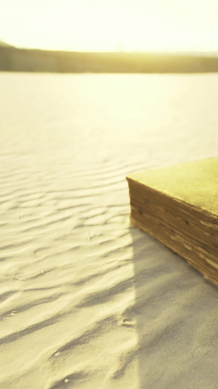 Old book resting on sand by a river during sunset with soft light shining