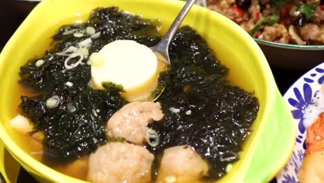 a spoon scoops silken tofu from a yellow bowl of clear broth with pork meatballs, seaweed, and scallions under bright, even lighting. close-up, steady camera