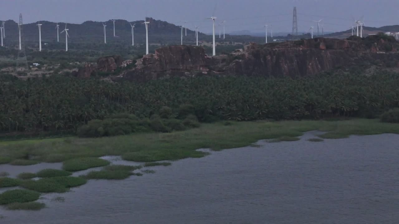 Scenic aerial footage showing wind turbines on hills during evening time, a lake with rocky terrain, and a vast mountain range under moody twilight clouds