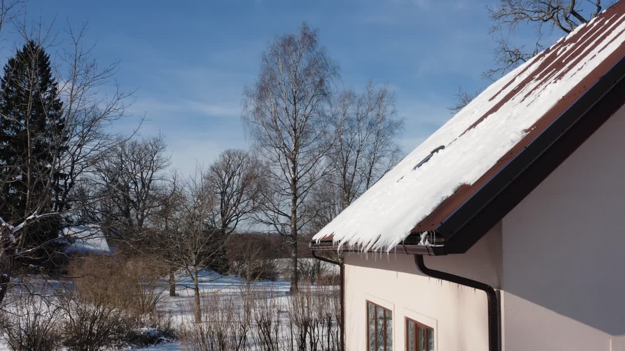 Snow falling of a home roof in countryside. Snow cascading from countryside home roof on a peaceful winter day. Serene rural winter scene.