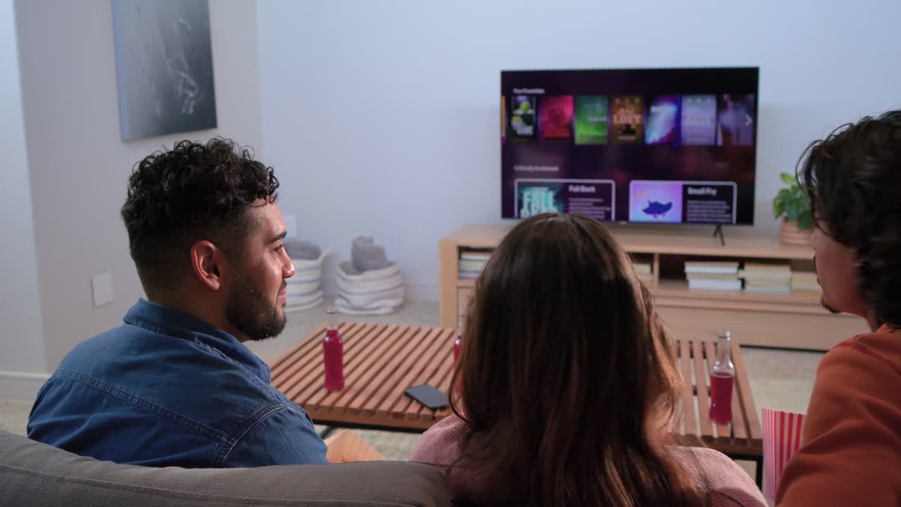 Różni znajomi wybierają zdalny i przeglądają menu przesyłania strumieniowego na telewizorze zamontowanym na ścianie, aby wybrać program