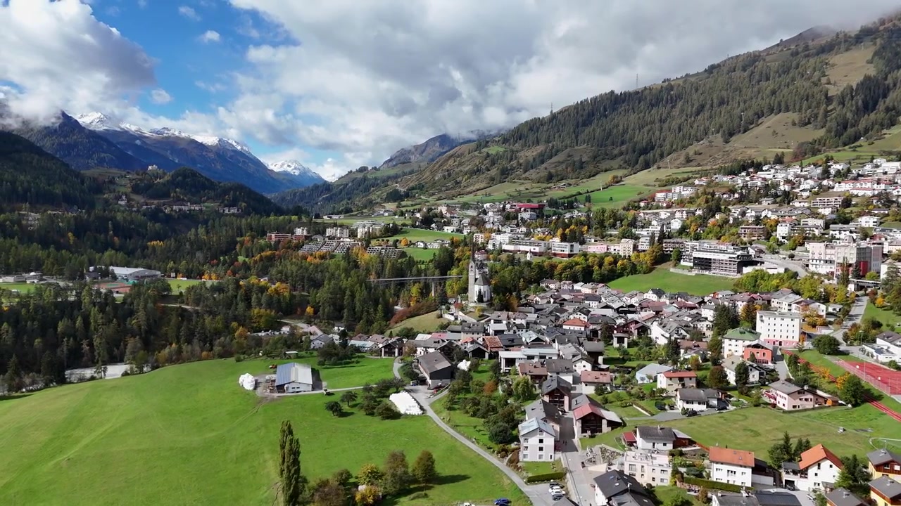Veduta aerea del villaggio di Scuol con case in una valle idilliaca della Svizzera. Erba verde con alberi colorati e montagne innevate sullo sfondo. Giornata di sole in autunno. Inquadratura ampia orbitale.