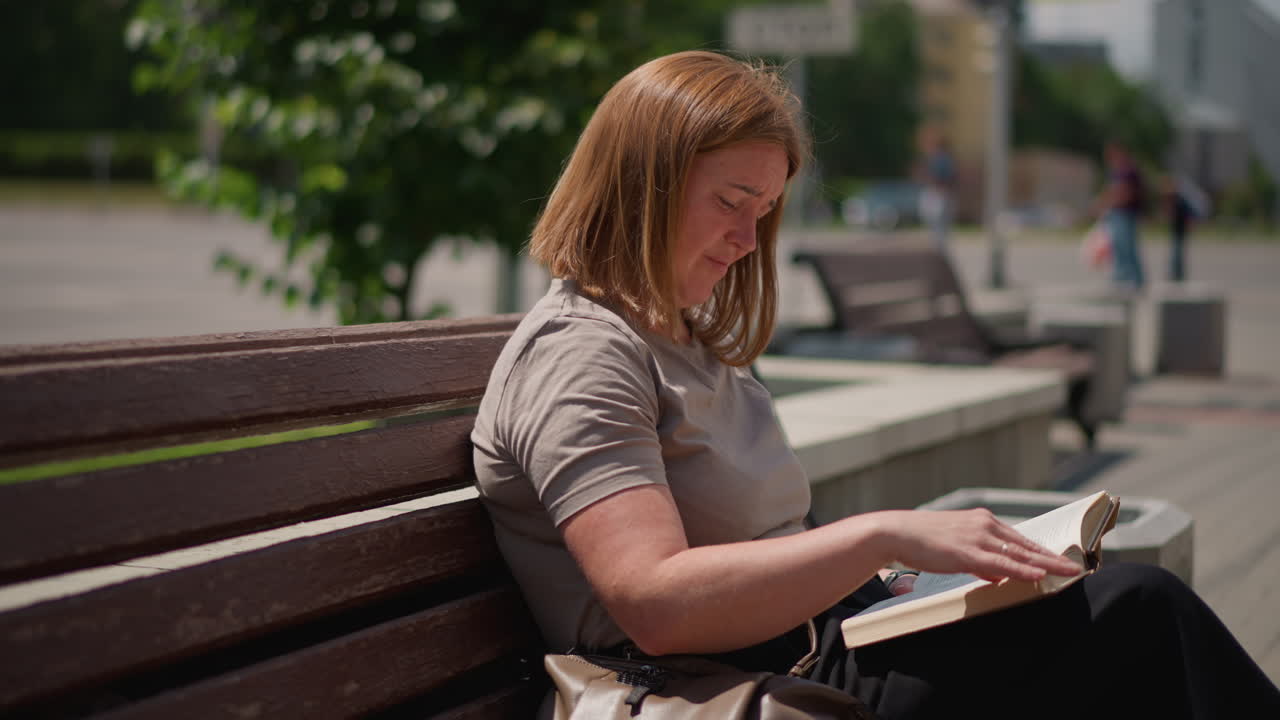 Woman sitting on wooden bench flipping page of open book under bright sunlight, focused expression and calm mood surrounded by greenery, soft breeze and gentle light