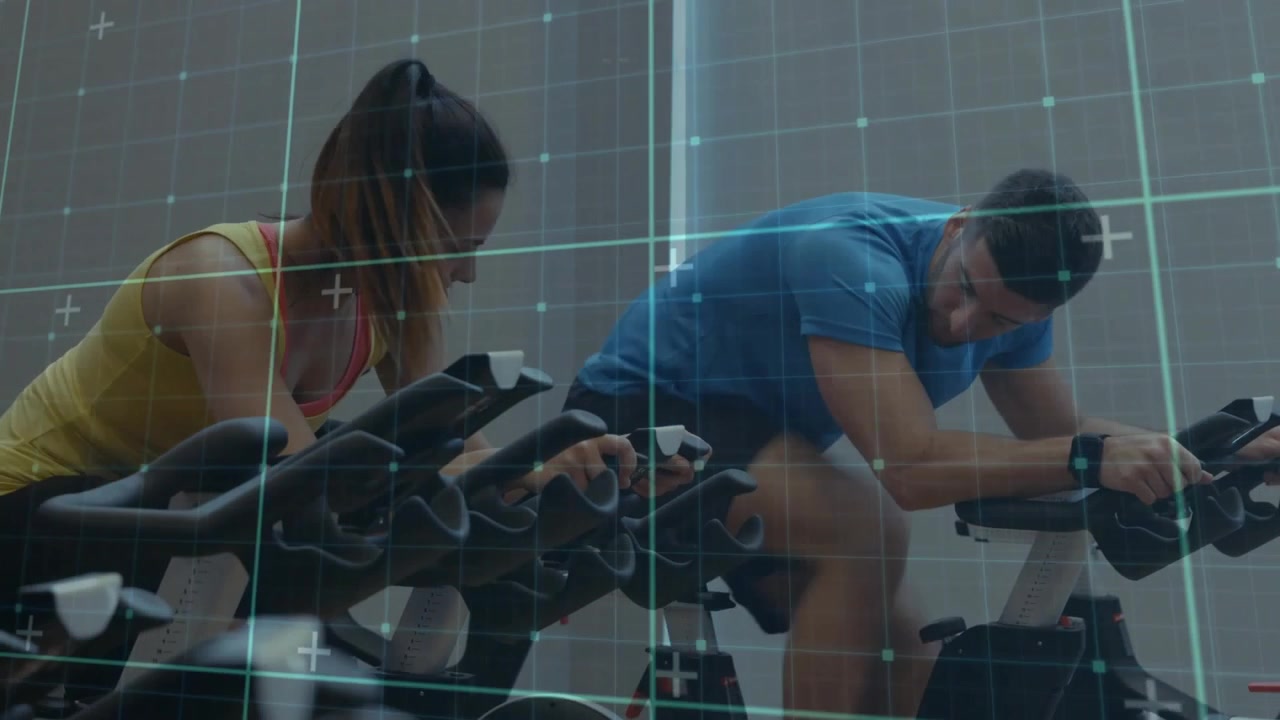 Vertical video: Pedaling woman in tank checking console prompt, man in blue pointing, wrist tracker