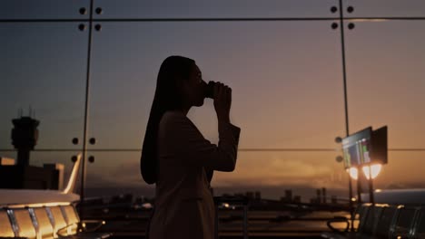 항공사 허브의 탑승 라운지에서 굴러가는 가방을 들고 비행기를 기다리는 동안 커피를 마시는 아시아 사업가, 비행기 터미널에서 비행기가 창밖에서 이륙합니다.