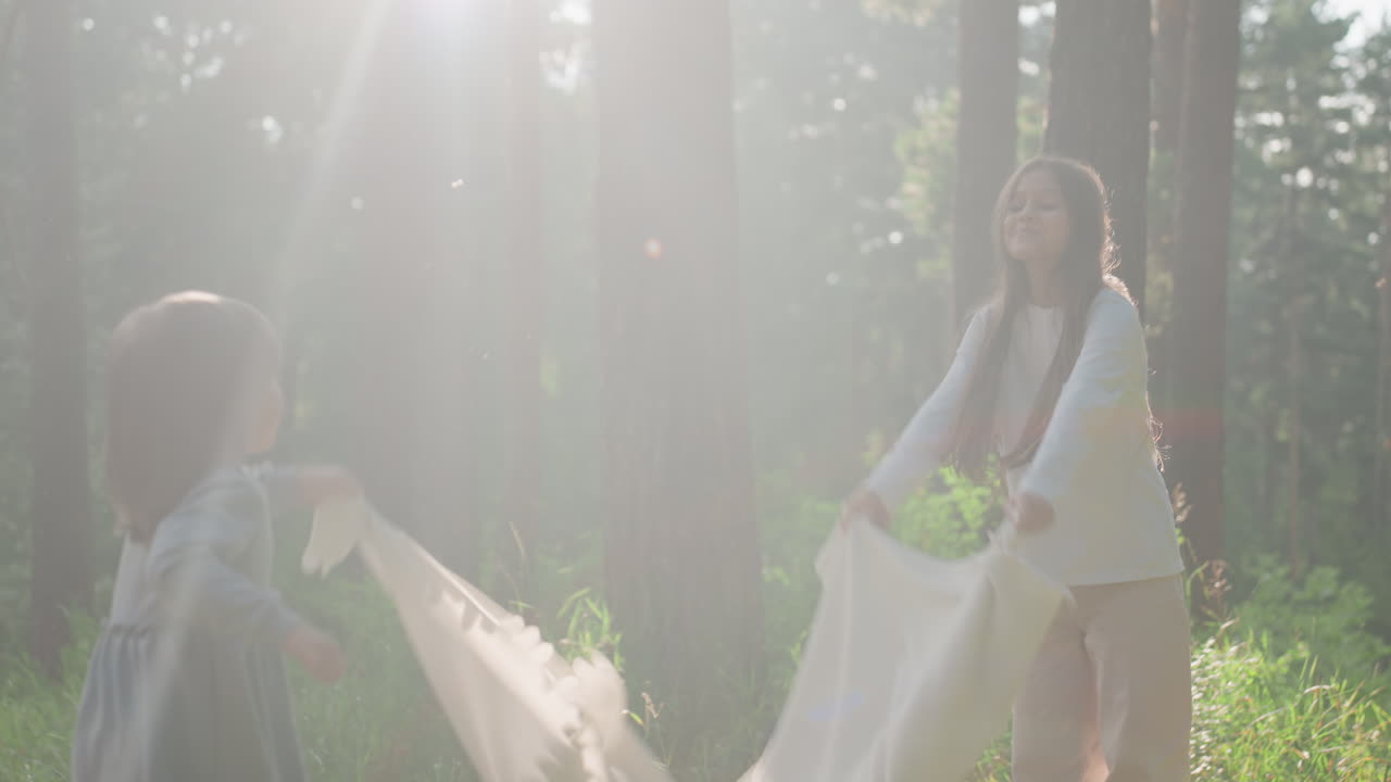 Playmates swinging white cloth playfully under sunlight in forest clearing, enjoying carefree outdoor fun together surrounded by trees, laughter, and summer warmth in peaceful natural atmosphere