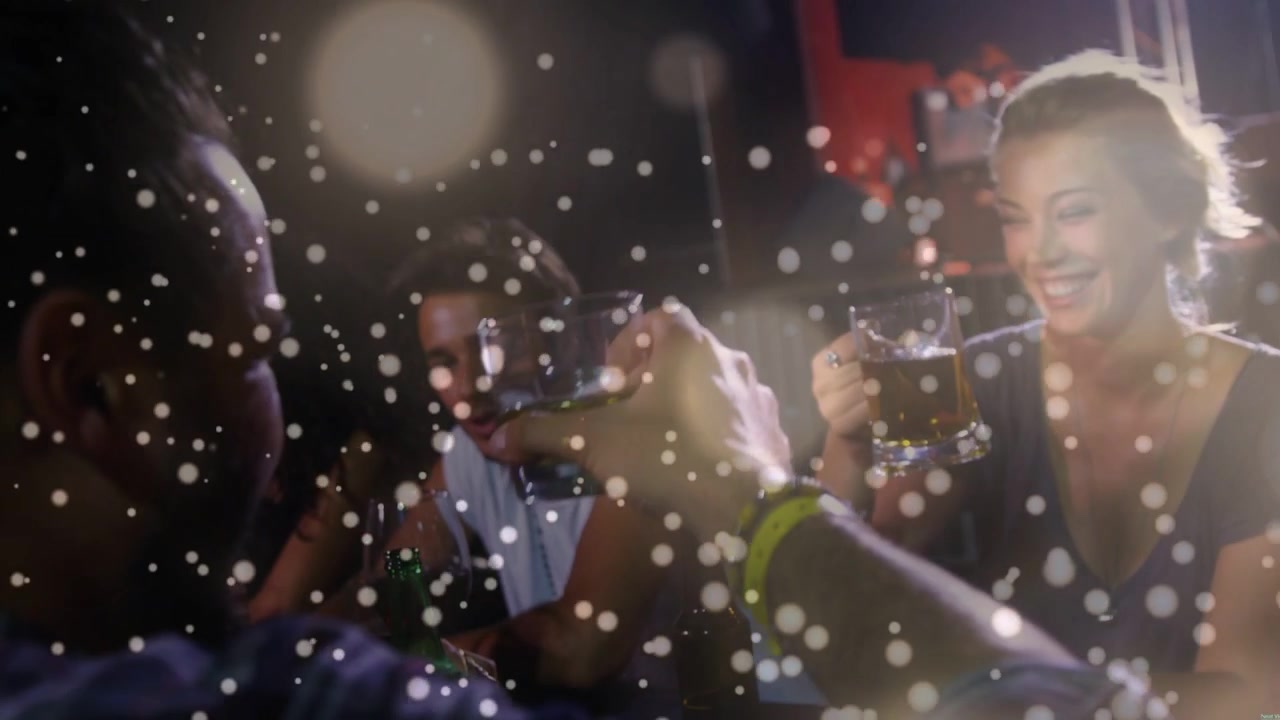 Mand hæver glas mod kvinde i bar, skåler med ølkrus, der klinger under bokeh-linseflare.