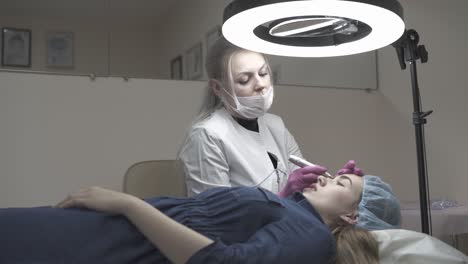 skilled woman in mask and coat applies permanent make-up