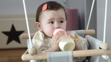 Süßes-Einjähriges-Mädchen,-Das-Eine-Flasche-Milch-Trinkt,-Während-Es-Auf-Einer-Babyschaukel-Drinnen-Sitzt---Nahaufnahme