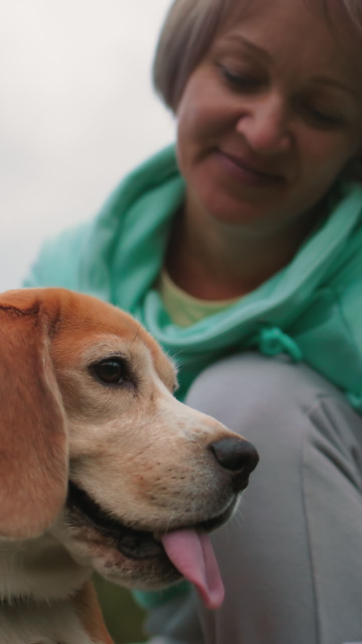 미소를 짓는 여성이 야외에서 비글 강아지를 무릎에 얹고 부드럽게 쓰다듬고 있어요. 청록색 후드티를 입었고, 하늘은 흐립니다. 얼굴을 클로즈업하여 따뜻하고 친밀한 유대감을 보여주며 편안한 오후, 부드러운 자연광, 공원 배경, 정서적 편안함이 느껴집니다.