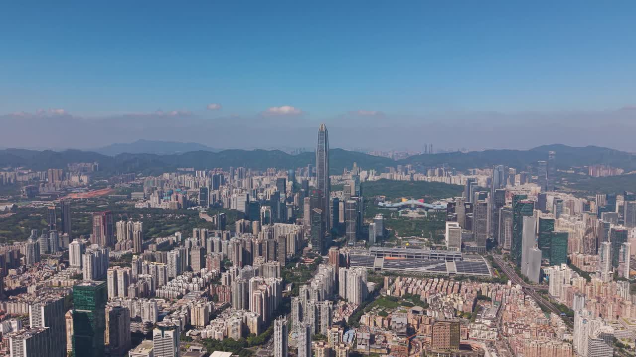 Wide drone shot of Shenzhen’s skyline featuring Ping An Finance Centre rising above dense urban districts. Modern high-rises, green park areas, and distant mountains under a clear blue sky. China