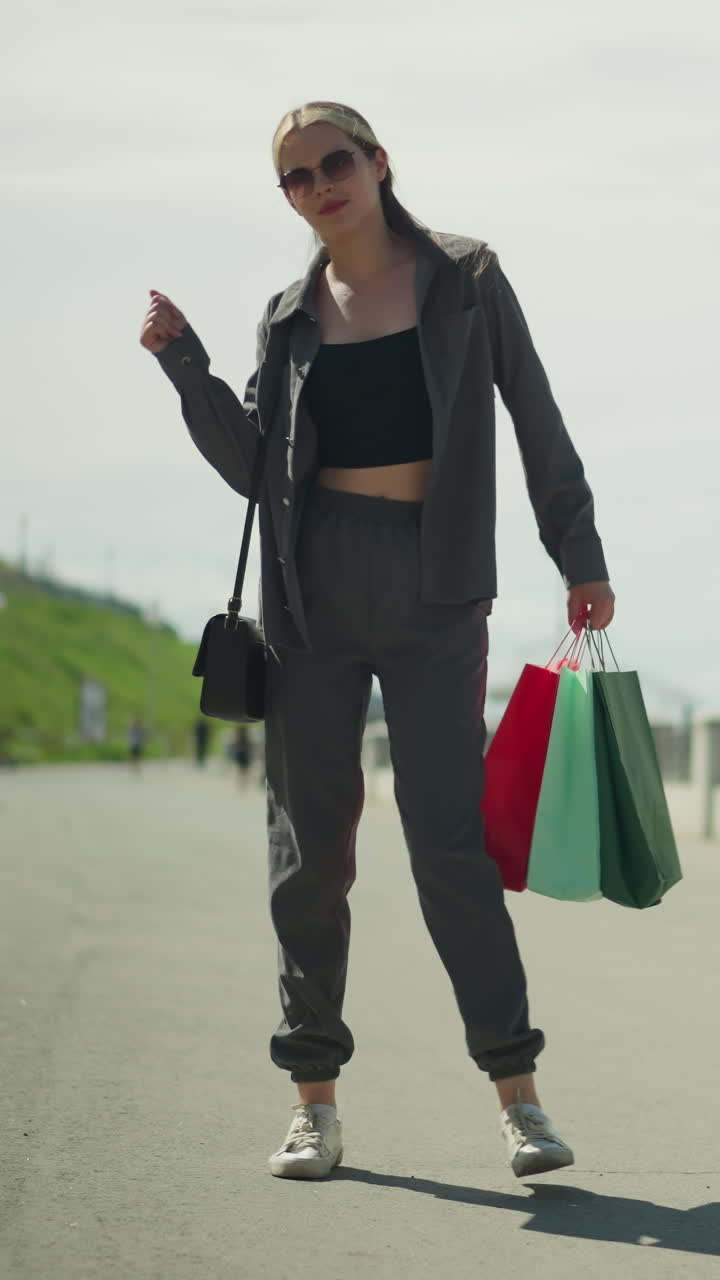 dama con camiseta negra bajo chaqueta gris baila alegremente, sosteniendo bolsas de compras rojas, menta y verdes en su mano derecha, su sombra es visible en el suelo, con personas caminando en el fondo