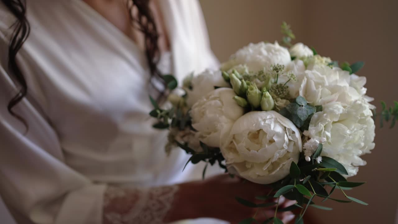 Primo piano della sposa in vestaglia di seta che tiene in mano un delicato bouquet da sposa bianco all'interno