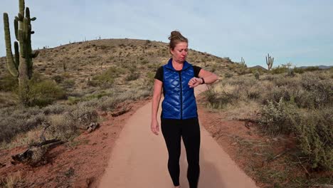 Mujer-Caminando-Y-Revisando-El-Reloj-Inteligente-En-Un-Sendero-Pavimentado-Del-Desierto