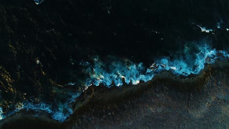 Aerial-top-down-perspective-as-whitewash-ocean-waves-crash-on-rocky-volcanic-shoreline,-curacao-drone