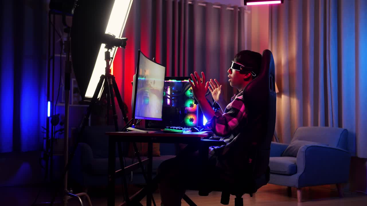 Side View Of Asian Boy Streamer Playing Game, Touching, And Swiping Experience Virtual Reality In Futuristic Goggles. Personal Computer On Desk Illuminated By Rgb Led Strip Light