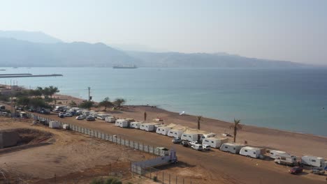 hadatiyim beach camping site with many caravan cars parked at the coastline on a sunny day, eilat city, israel - aerial dolly right, revealing self-elevating platform for shipment in shallow waters