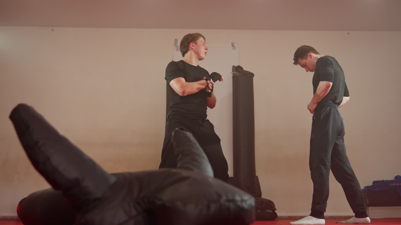 Wrestlers in gym, one athlete adjusting waistband while another lift him up and throw on red mat with punching bags and equipment visible in background