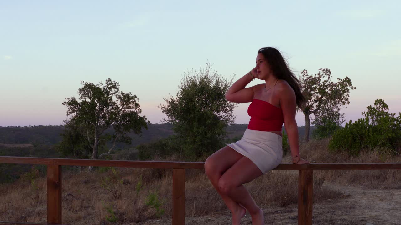 una joven modelo femenina se sienta en una valla de madera durante la puesta del sol, acariciando su largo cabello oscuro