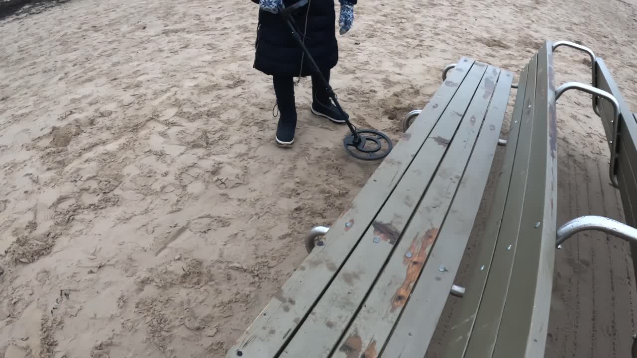 En person bruger en metaldetektor nær en vejrslidt strandbænk og scanner sandet ved siden af siddeområdet for mistede genstande på en kold kystdag