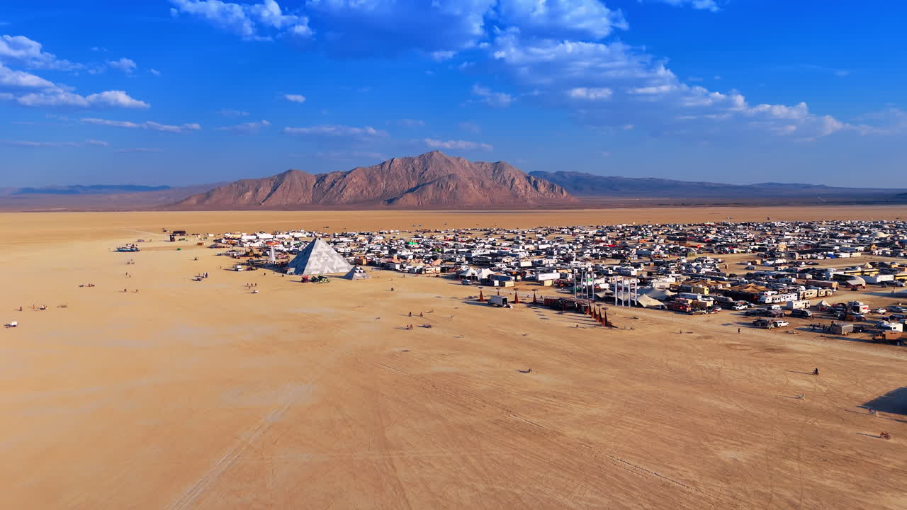 Kalifornia, USA, 1. heinäkuuta 2025: Suuri pyramidimainen rakennelma ja leiri laajassa aavikkomaisemassa. Ilmakuva geometrisesta pyramidiasennelmasta tilapäisen matkailuautokaupungin vieressä sinisen taivaan alla.