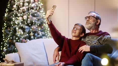 Porträt-Eines-Glücklich-Lächelnden-Alten-Opas-Mit-Einem-Jugendlichen-Enkelkind,-Das-In-Einem-Dekorierten-Zimmer-Sitzt-Und-Selfie-fotos-Auf-Dem-Smartphone-Zu-Hause-In-Der-Nähe-Des-Glühenden-Neujahrsbaums-Macht