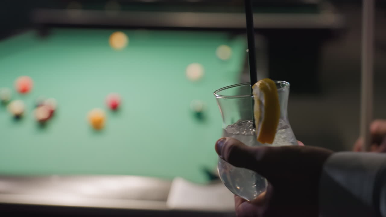 Close-up of person's hand holding glass of lemon drink with black straw. Ice cubes and lemon slice add refreshing detail. Blurred pool table with colorful balls creates a dynamic and relaxing ambiance