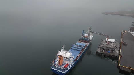 Entladen-Von-Fischen-Aus-Einem-Frachtschiff-An-Einem-Nebligen-Tag-In-Faskrudsfjördur,-Ostisland---Drohnenaufnahme-Aus-Der-Luft