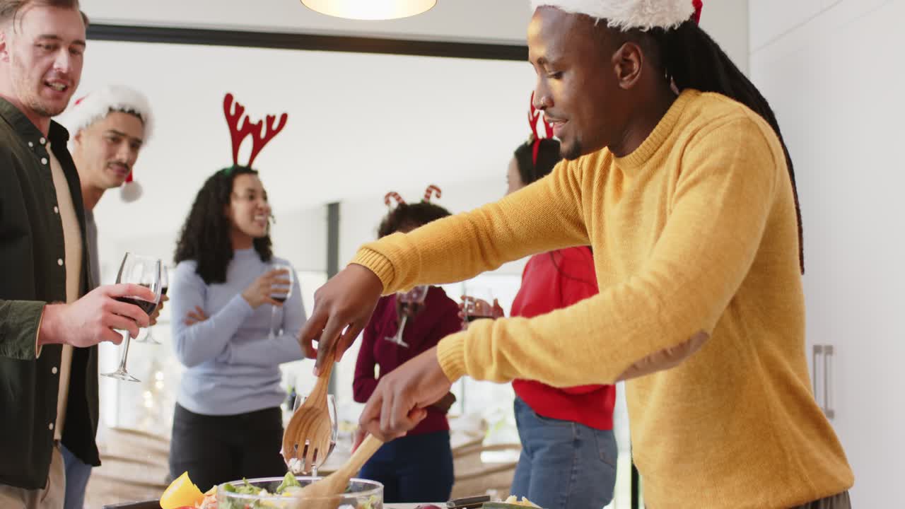 Amici di diverse etnie con cappelli da Babbo Natale, bicchieri in mano, che incoraggiano un uomo di colore ad affettare cetrioli e mescolare l'insalata.