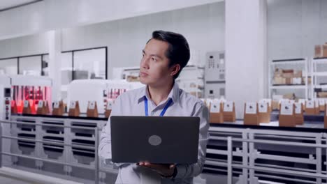 asian male professional worker standing with his laptop with conveyor for packaging coffee beans in bags at coffee factory, he observes by looking around before concentrating with his laptop