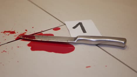 a hand wearing a glove, putting a card with a number near a knife soaked in blood on the floor