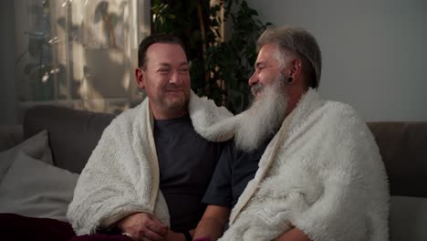 un feliz hombre moreno con una camiseta gris envuelto en una manta blanca de lana se sienta en el sofá y habla con su novio mayor con cabello gris y una exuberante barba blanca en un apartamento moderno. pareja lgbt se comunica y pasa tiempo juntos