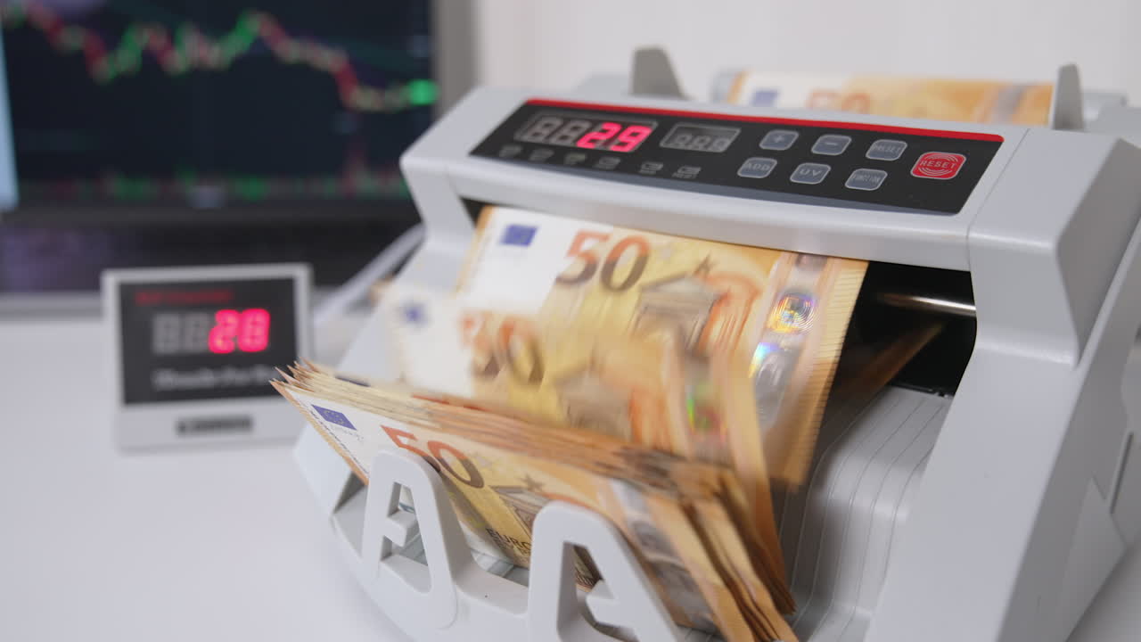 Money counting machine with a wad of fifty euro banknotes. Close up. Laptop opened at backdrop in blur.