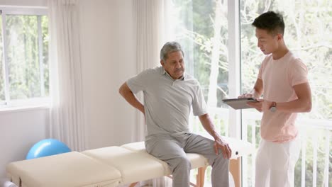 diverse male physiotherapist with tablet examinig senior male patient, in slow motion