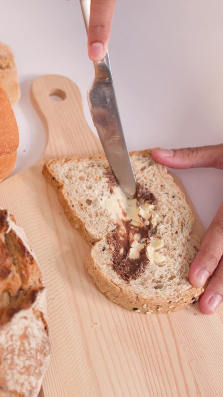 Close-up overhead shot of hands spreading creamy nut butter and chocolate onto multigrain bread.