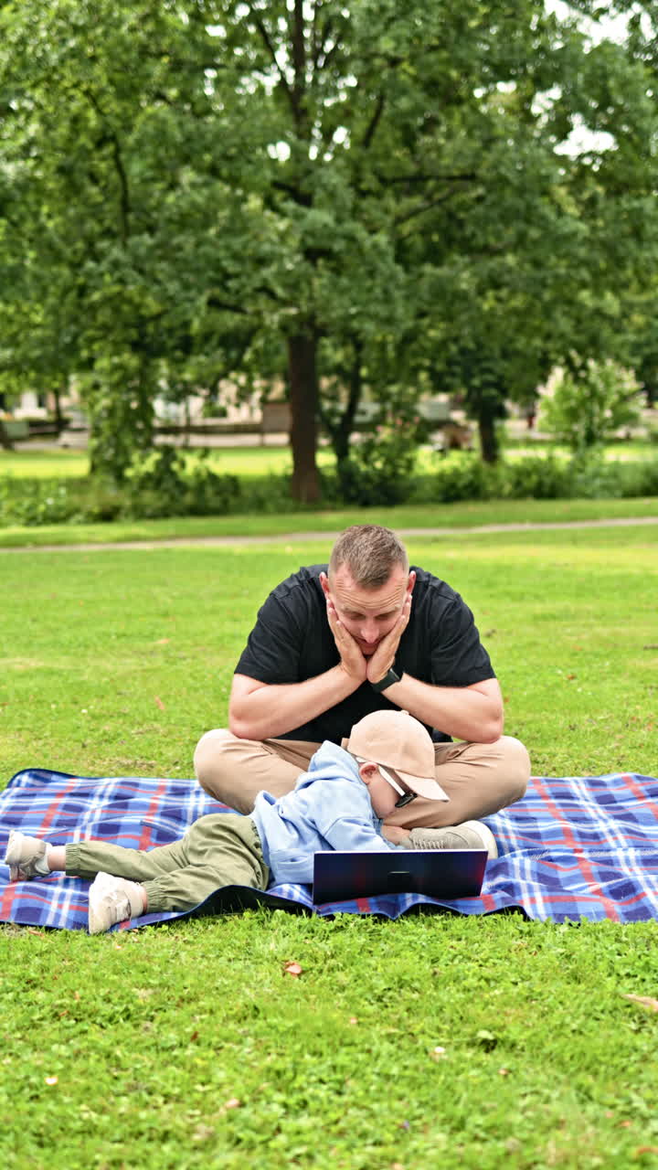 En ung far sidder på græsset med sin bærbare computer, mens hans lille søn leger i nærheden. En mand, der arbejder hjemmefra, sidder i parken, mens han er på tur med sin søn. Freelance-far og søn i naturen.