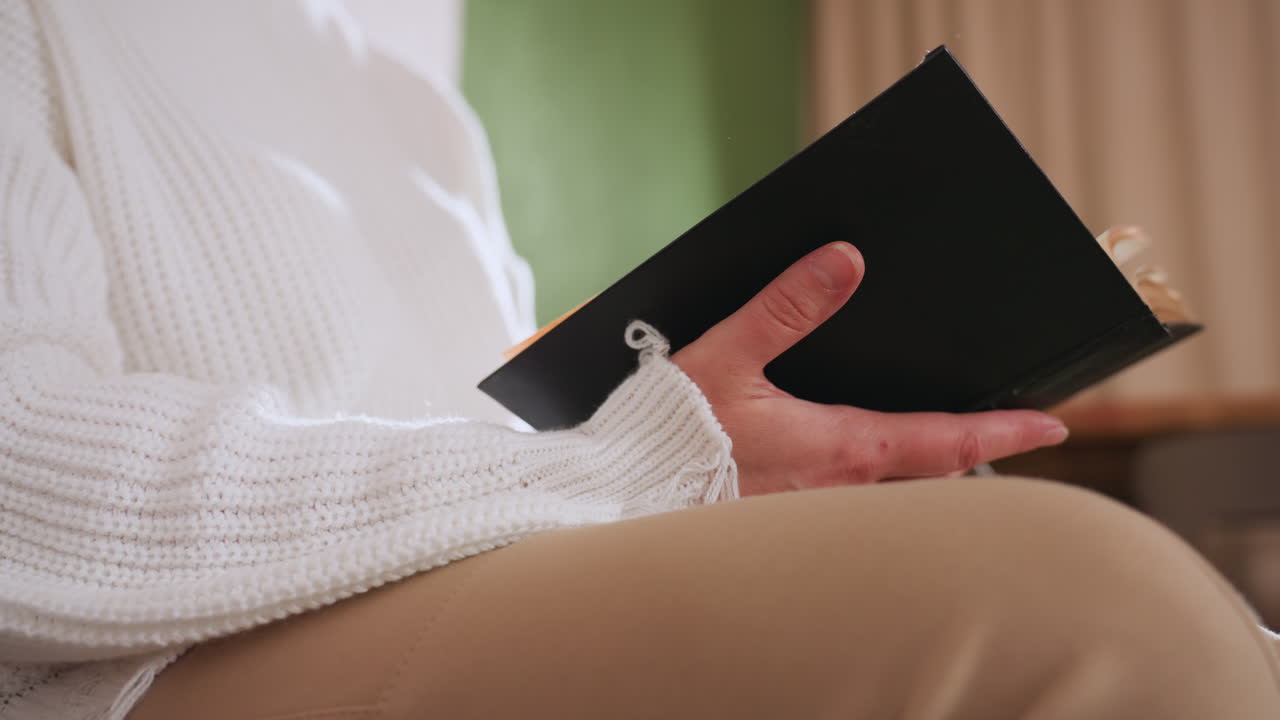 Lower view of mindful guest seated flipping pages of book with gentle hands, dressed in white sweater and beige pants, capturing quiet indoor ambiance and subtle reflective mood in soft lighting