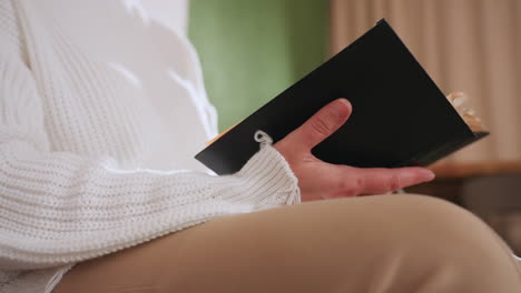 lower view of mindful guest seated flipping pages of book with gentle hands, dressed in white sweater and beige pants, capturing quiet indoor ambiance and subtle reflective mood in soft lighting