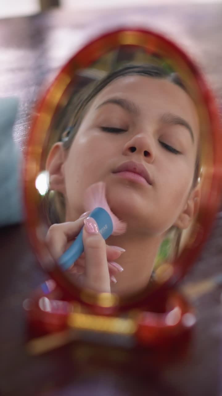 Primer plano del retrato de una adolescente mirándose al espejo y maquillándose con una brocha suave en la mejilla, tocador enmarcado por un espejo rojo ornamentado, luz natural y bokeh suave, expresión concentrada practicando la técnica de maquillaje